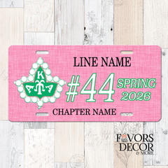 A pink novelty license plate with customizable text and a green and white floral emblem, displayed on a light wood plank background. The plate includes placeholder text for 'LINE NAME', Line number, Crossing Season & Year, and 'CHAPTER NAME', with a 'Favors Decor & More' logo in the bottom right corner.