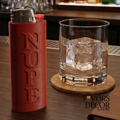 A crimson lighter sleeve with the word NUPE embossed vertically, shown next to a clear rocks glass with an ice cube on a wooden coaster at a bar. A 'Favors Decor & More' logo is visible in the lower right.