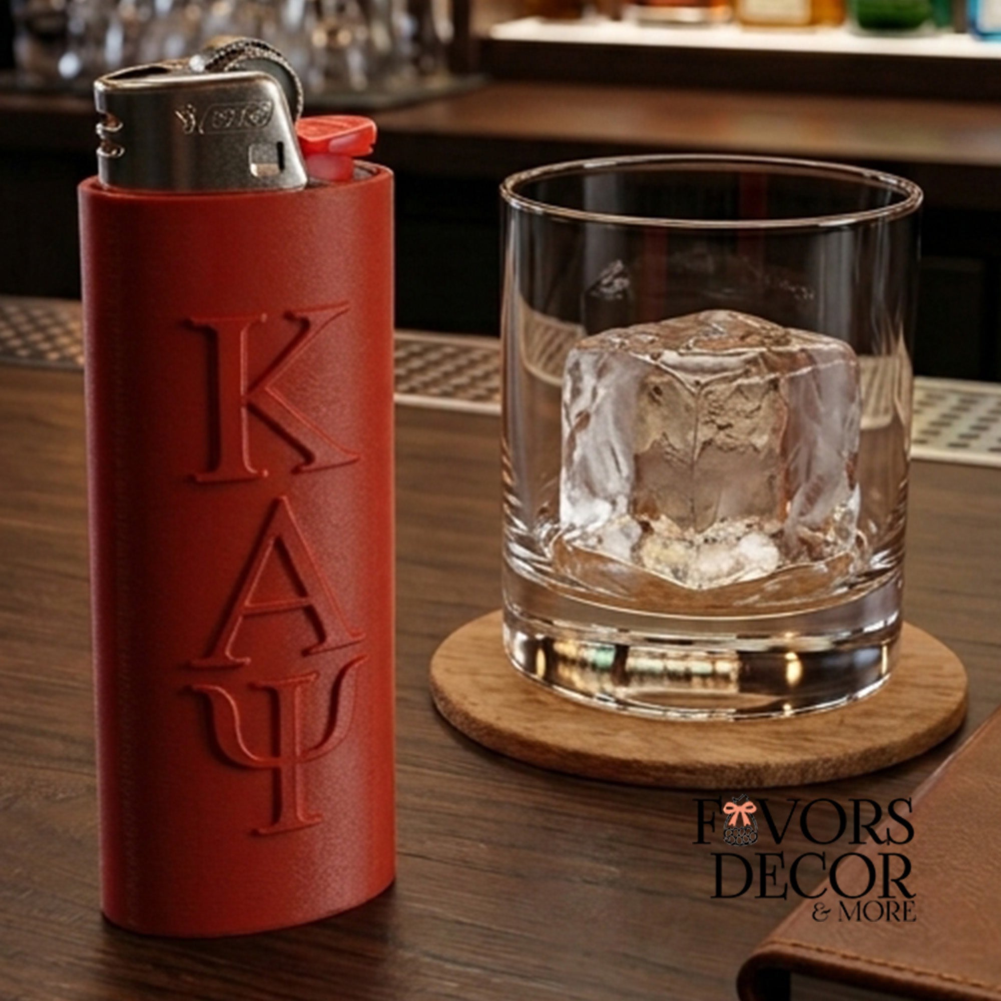 A crimson lighter sleeve with the word Kappa Alpha Phi greek letters embossed vertically, shown next to a clear rocks glass with an ice cube on a wooden coaster at a bar. A 'Favors Decor & More' logo is visible in the lower right.