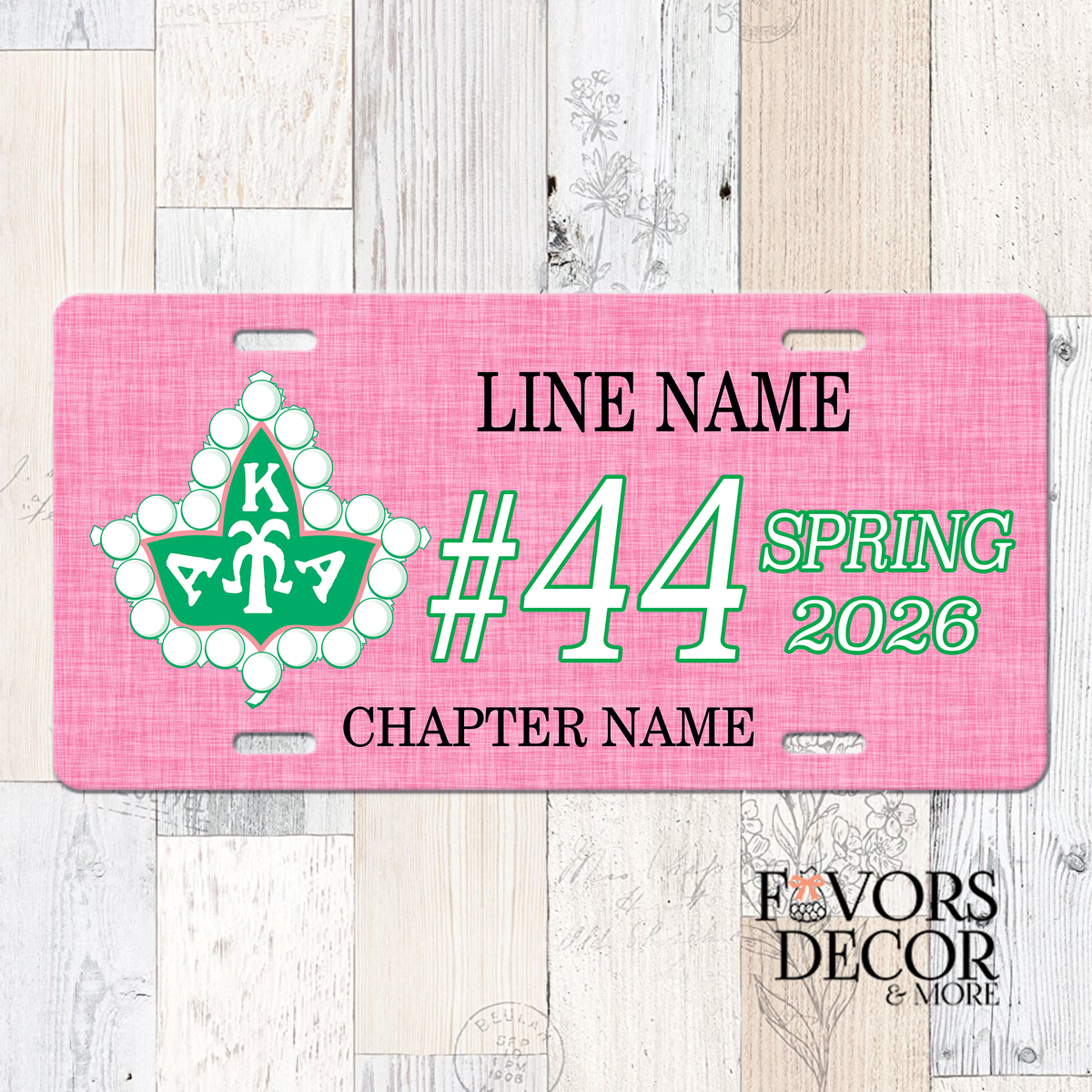 A pink novelty license plate with customizable text and a green and white floral emblem, displayed on a light wood plank background. The plate includes placeholder text for 'LINE NAME', Line number, Crossing Season & Year, and 'CHAPTER NAME', with a 'Favors Decor & More' logo in the bottom right corner.