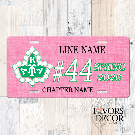 A pink novelty license plate with customizable text and a green and white floral emblem, displayed on a light wood plank background. The plate includes placeholder text for 'LINE NAME', Line number, Crossing Season & Year, and 'CHAPTER NAME', with a 'Favors Decor & More' logo in the bottom right corner.